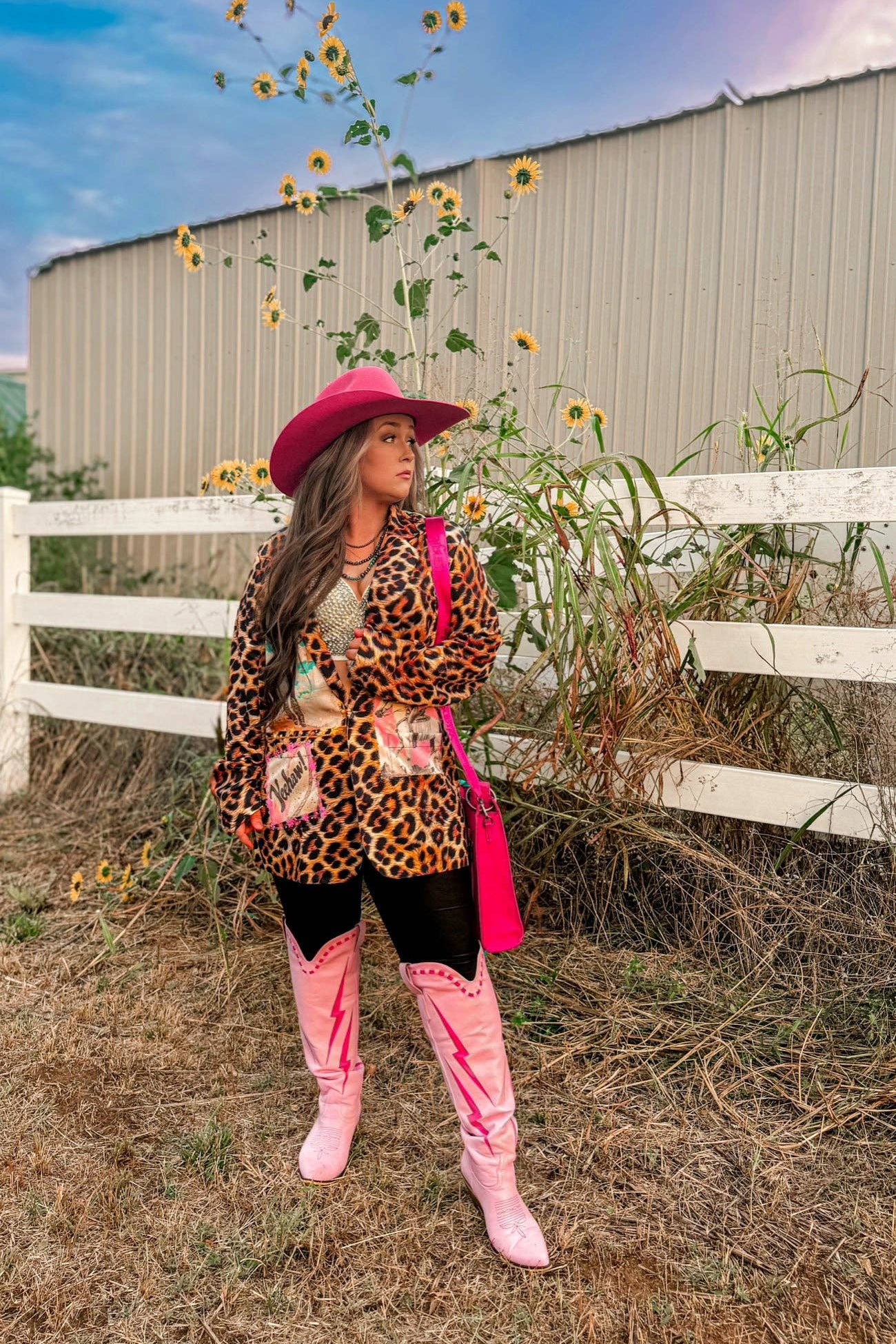 Person wearing a leopard print jacket, pink hat, and boots standing in a sunflower field with a white fence in the background.