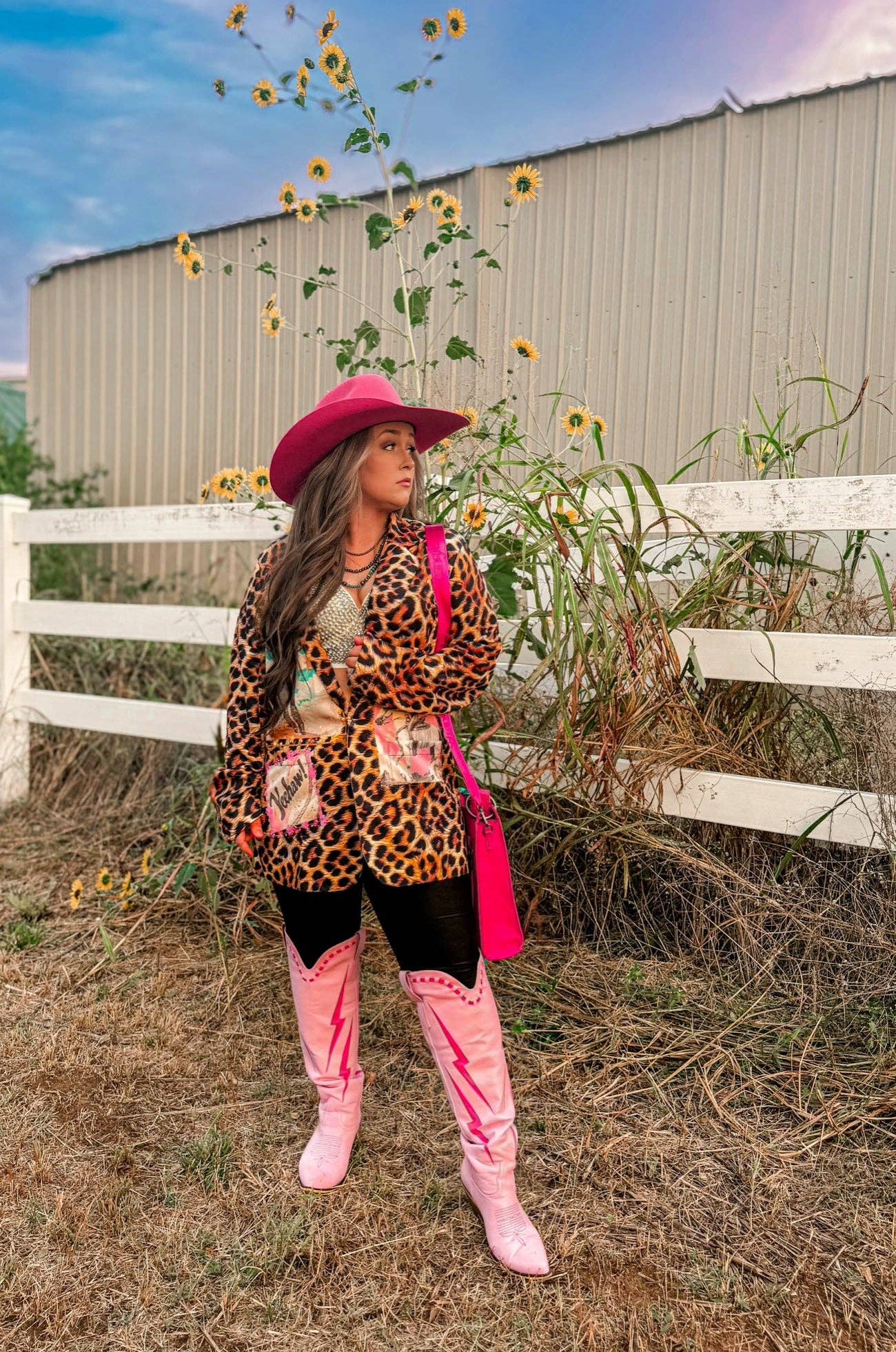 Person wearing a leopard print jacket, pink hat, and boots standing in a sunflower field with a white fence in the background.