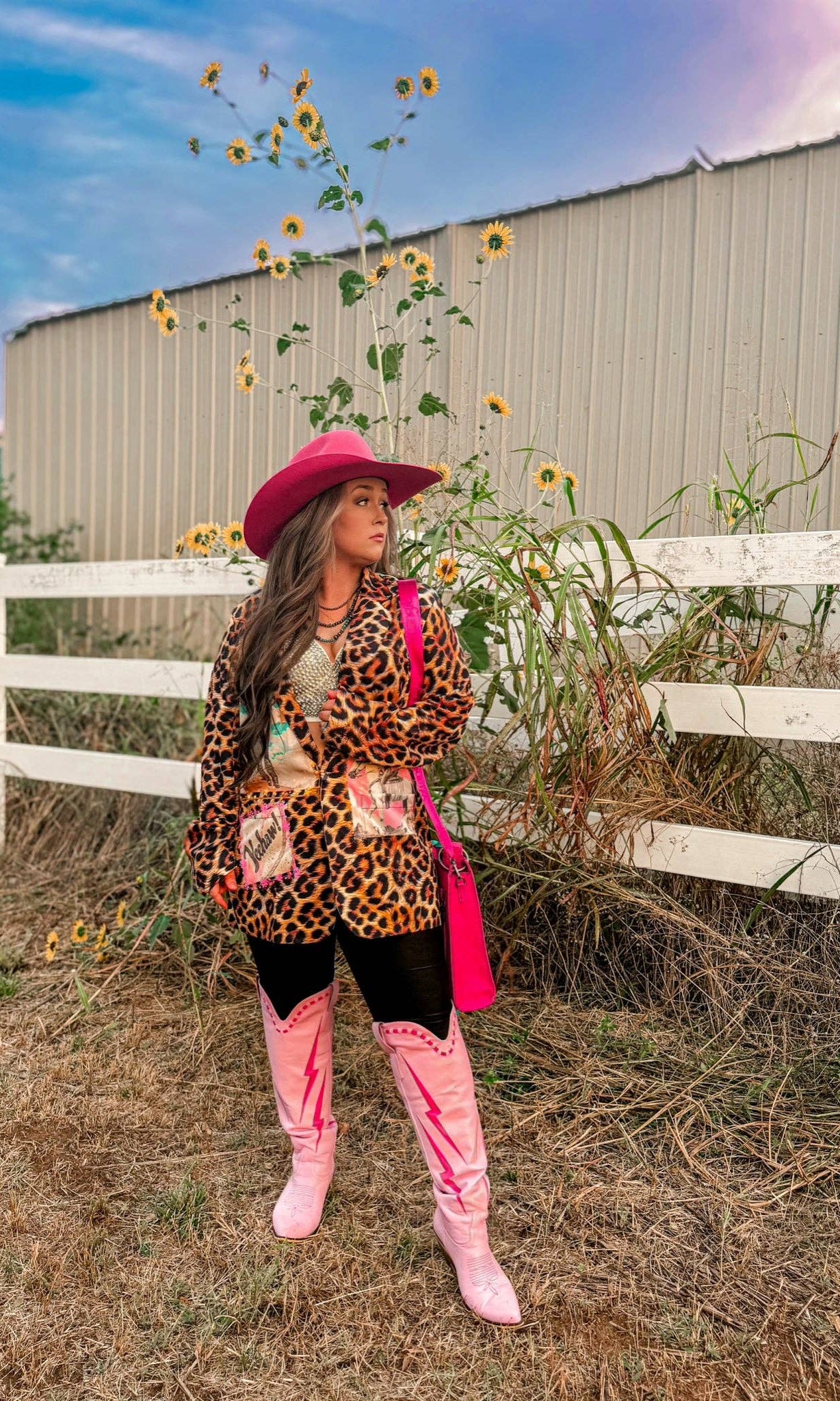 Person wearing a leopard print jacket, pink hat, and boots standing in a sunflower field with a white fence in the background.