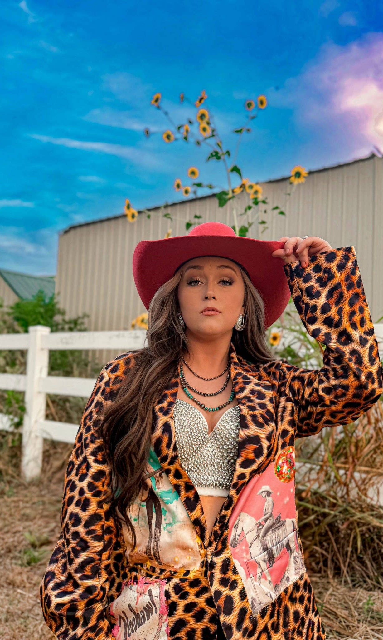 Woman in a leopard print jacket and red hat holding sunflowers against a blue sky.