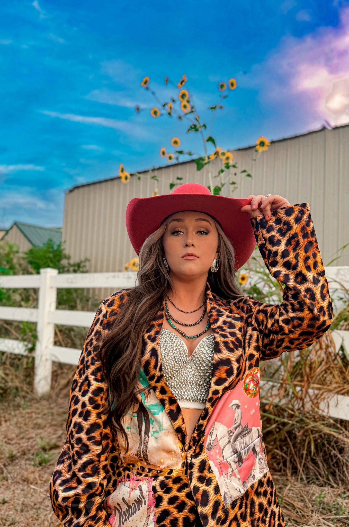 Woman in a leopard print jacket and red hat holding sunflowers against a blue sky.