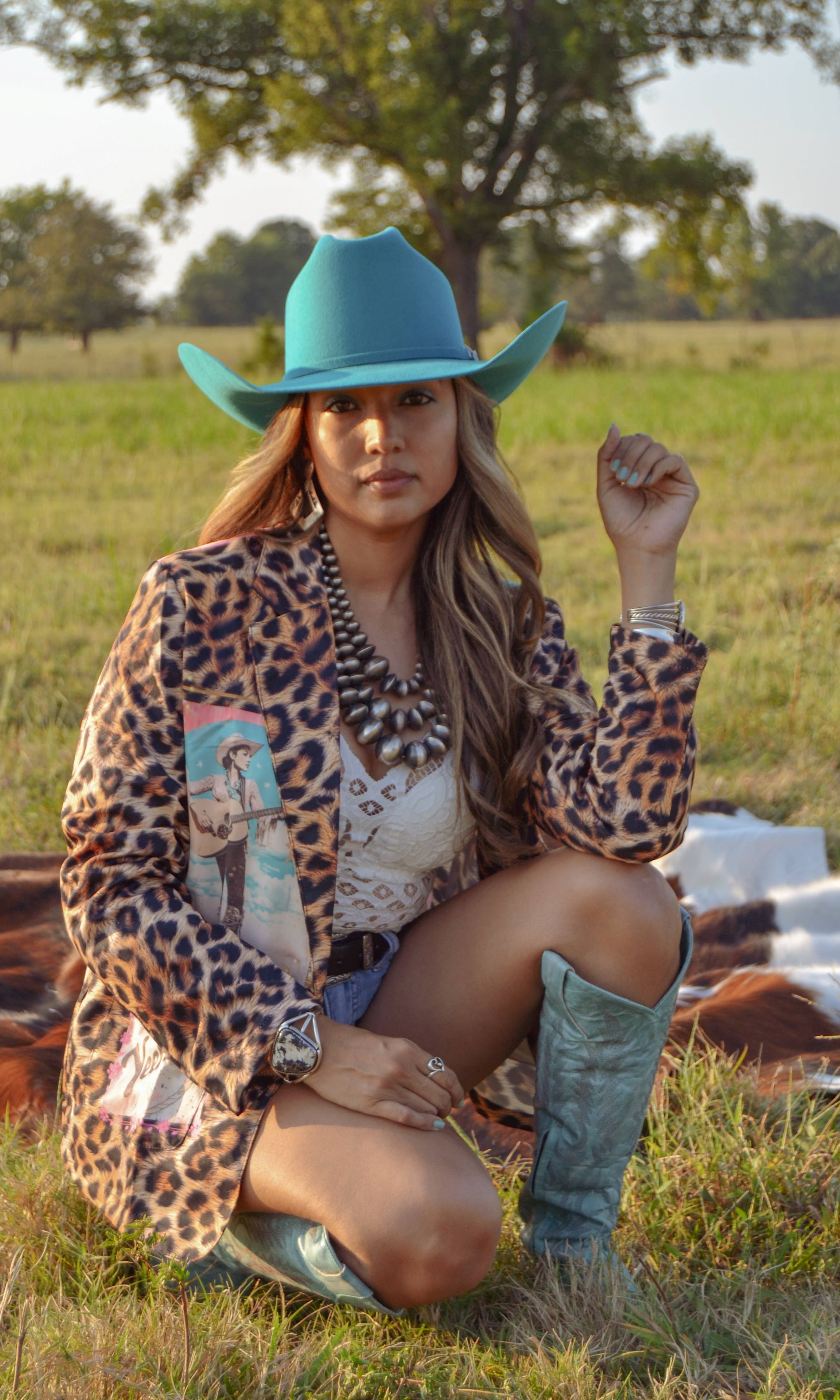 Woman in a leopard print jacket and blue cowboy hat sitting in a field.