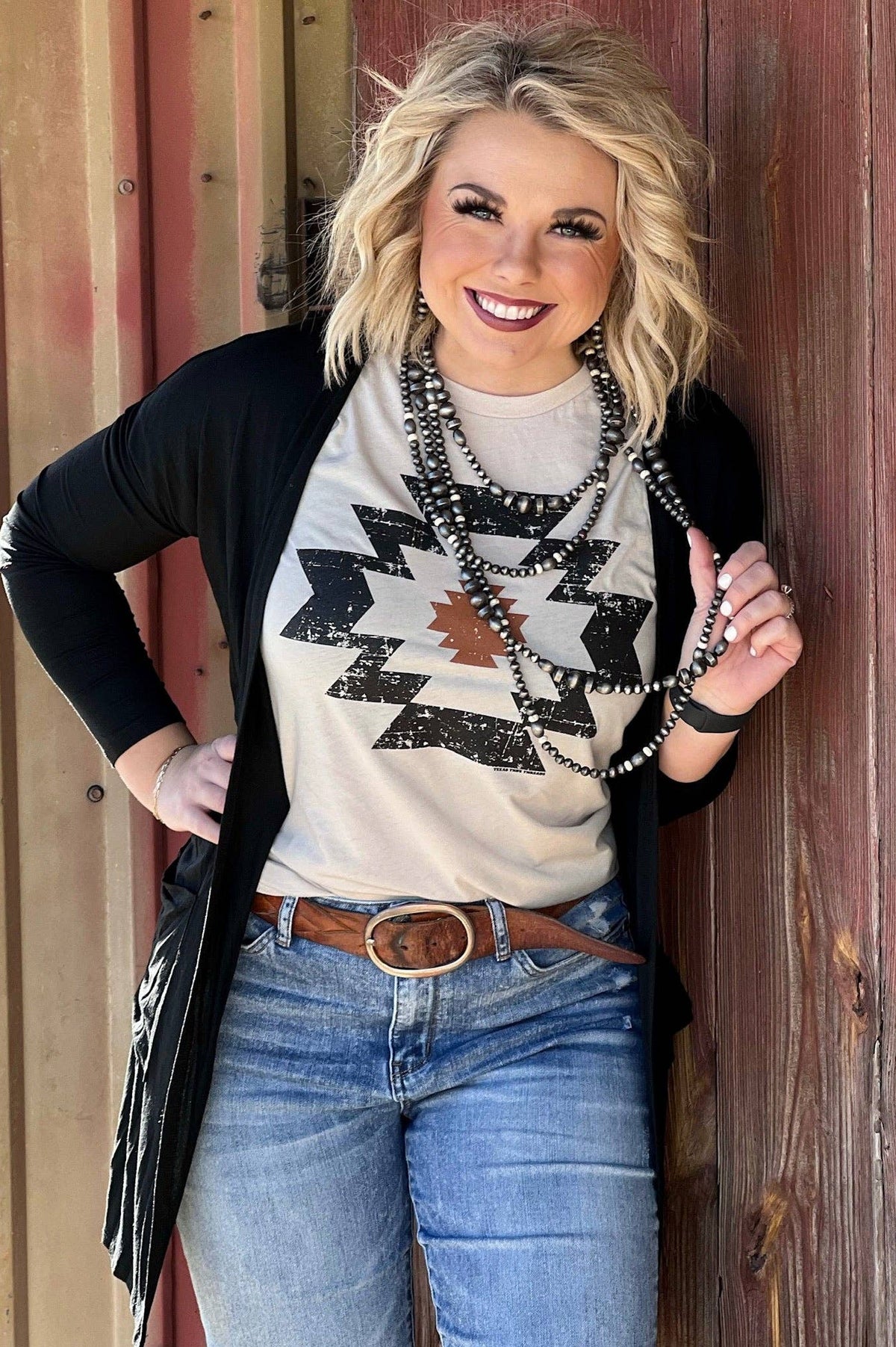 Woman wearing a graphic t-shirt, black cardigan, and blue jeans with a wooden background