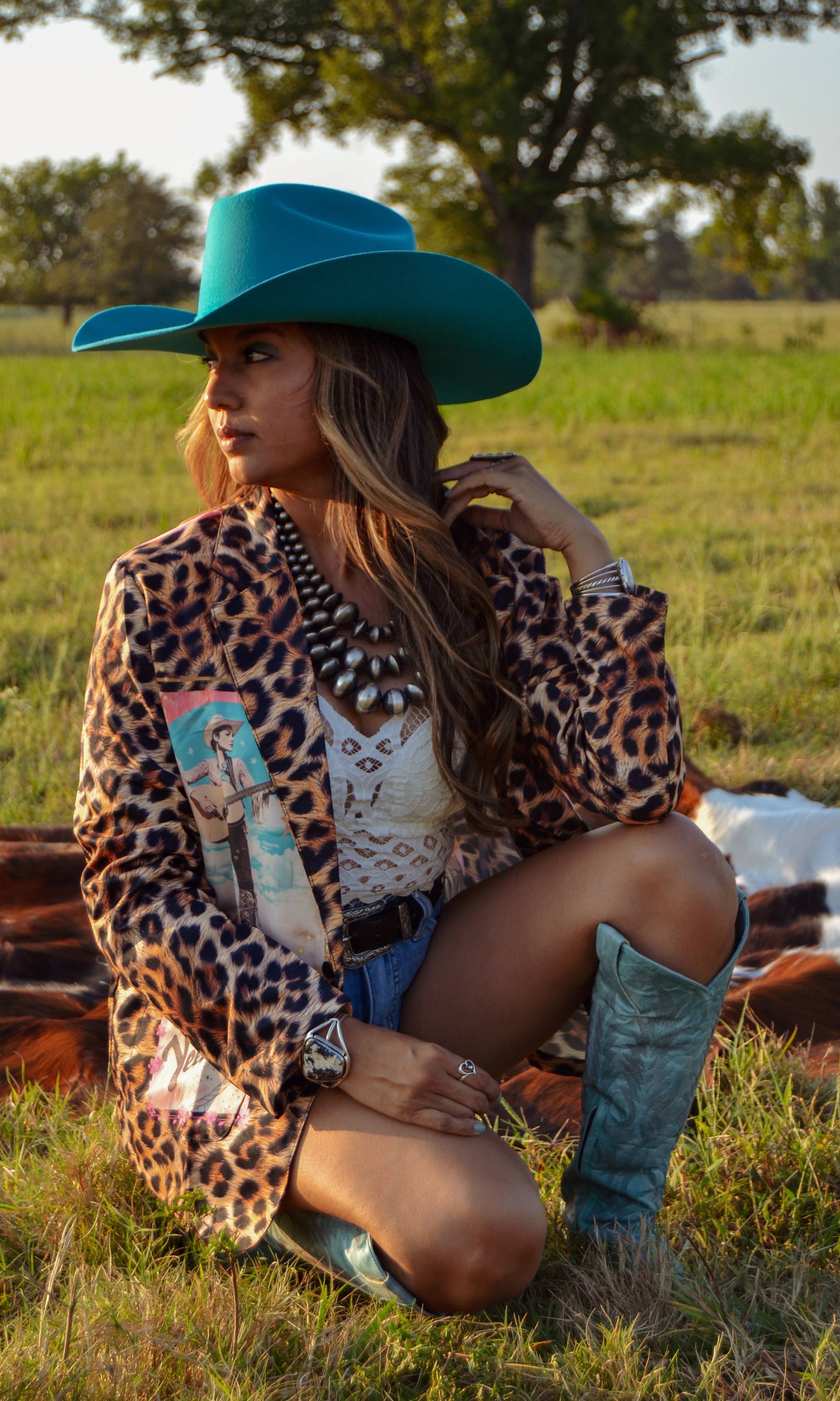 Woman in a leopard print jacket and blue cowboy hat sitting in a field.
