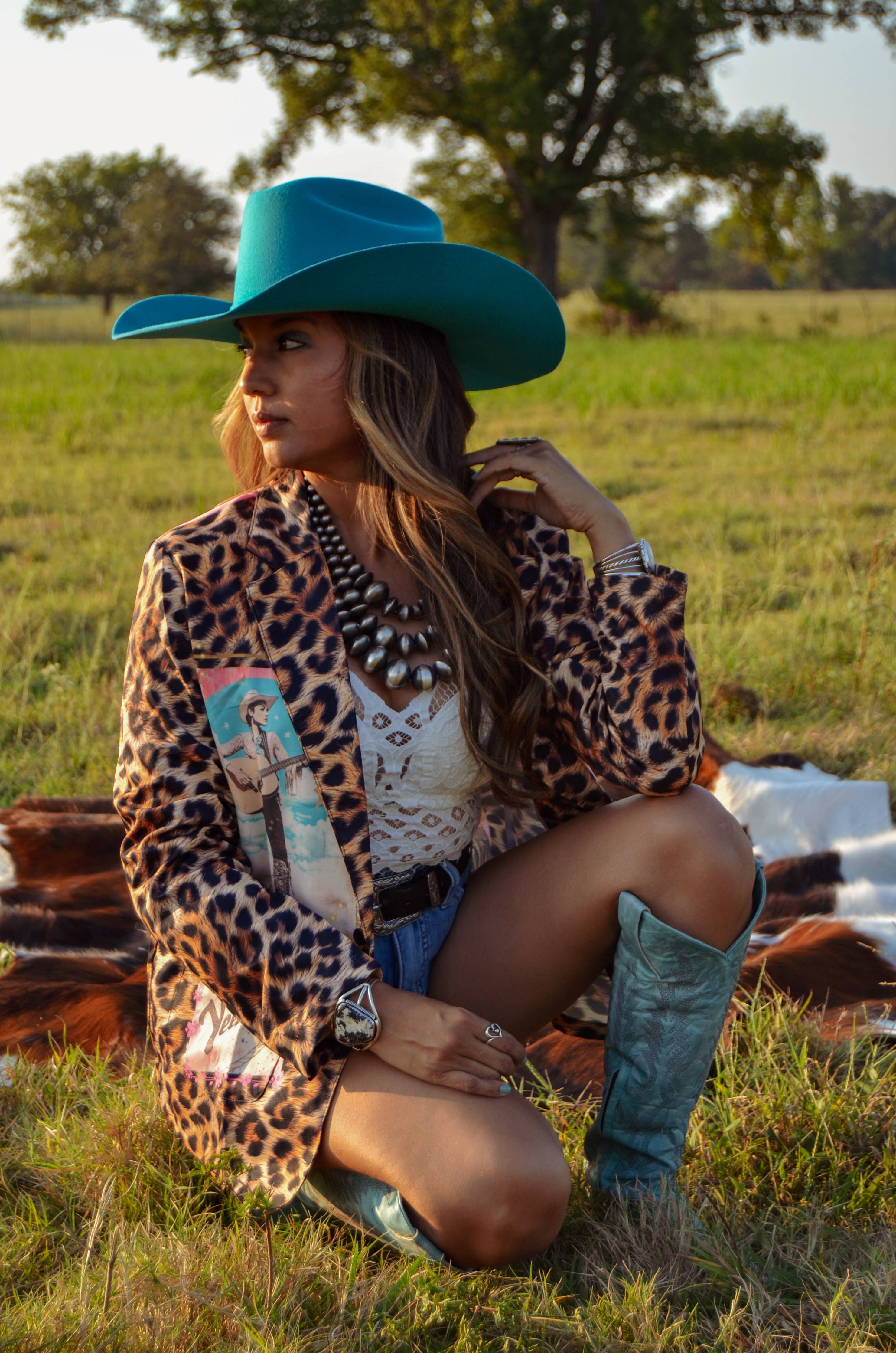 Woman in a leopard print jacket and blue cowboy hat sitting in a field.