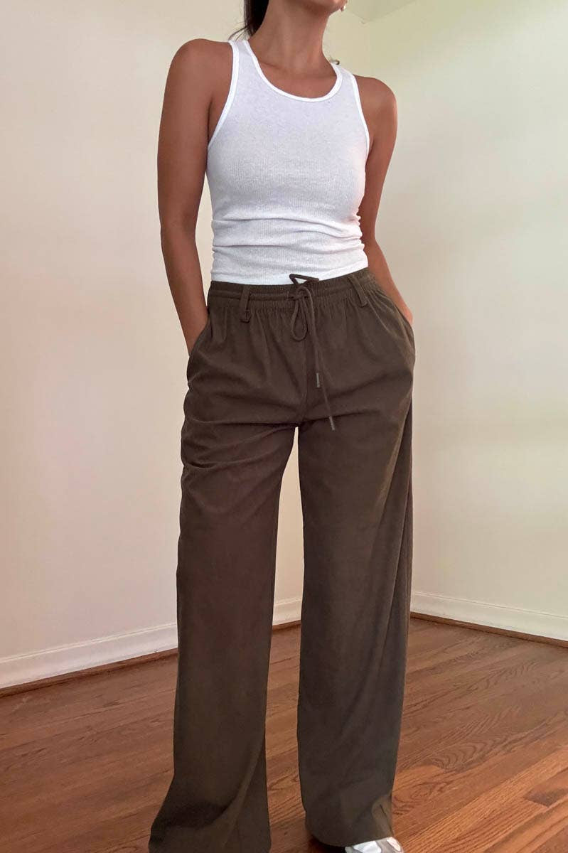 Person wearing a white tank top and brown pants in a room with a wooden floor.