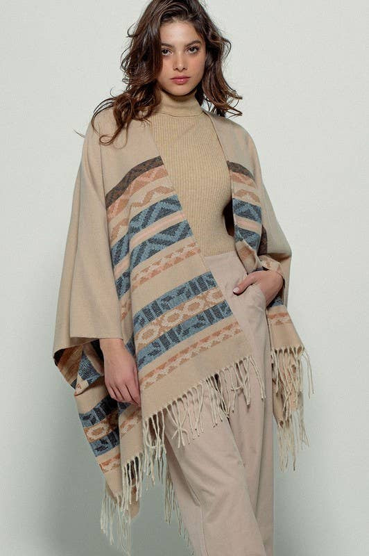 Woman wearing a beige poncho with a Tan background and light blue, brown, light rust, and grey aztec pattern design in stripes with tassel trim along bottom hem. Light weight fabric