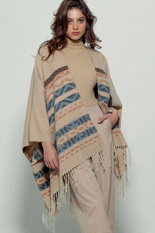 Woman wearing a beige poncho with a Tan background and light blue, brown, light rust, and grey aztec pattern design in stripes with tassel trim along bottom hem. Light weight fabric