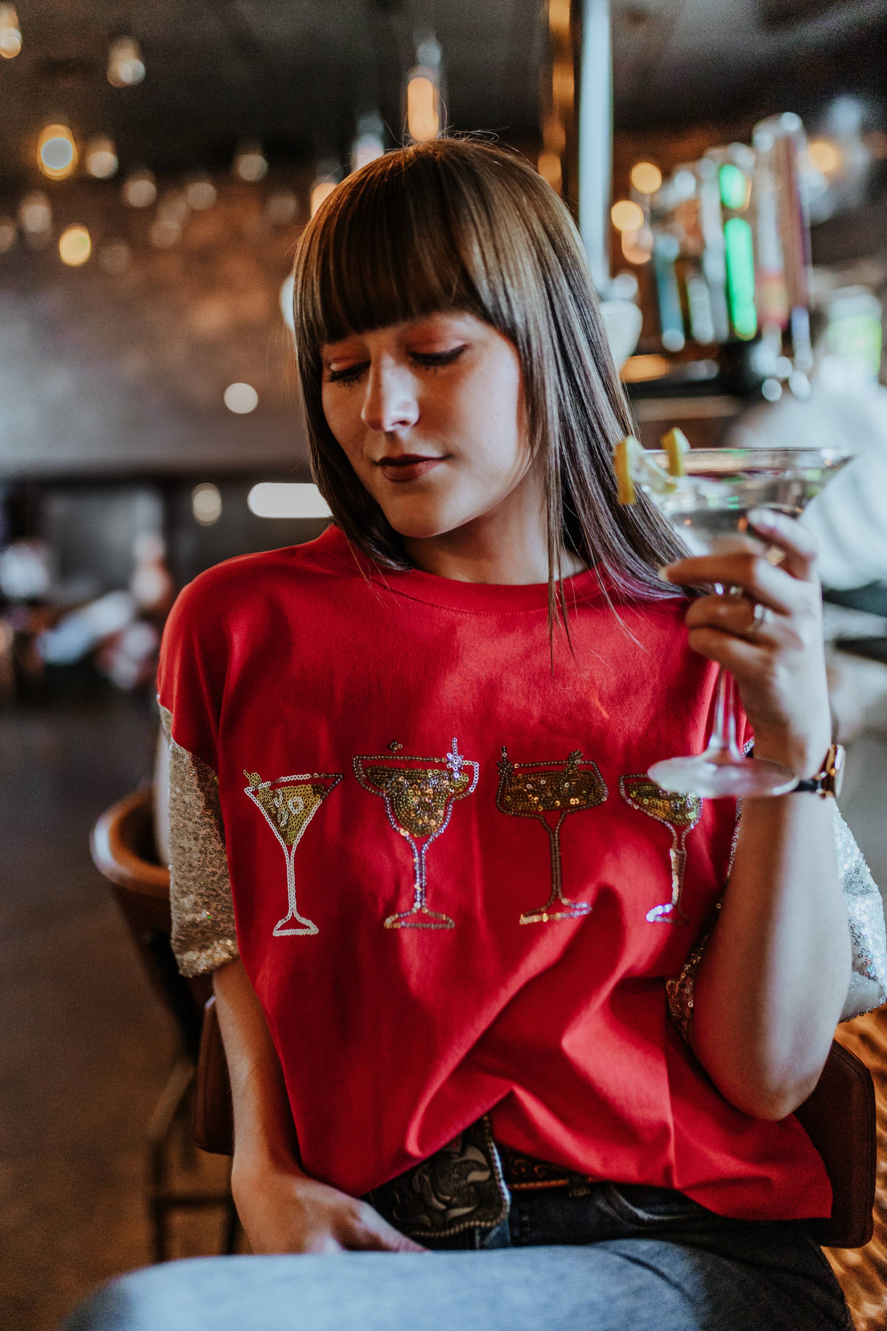 Woman in a red t-shirt with cocktail graphics holding a martini glass in a bar setting.