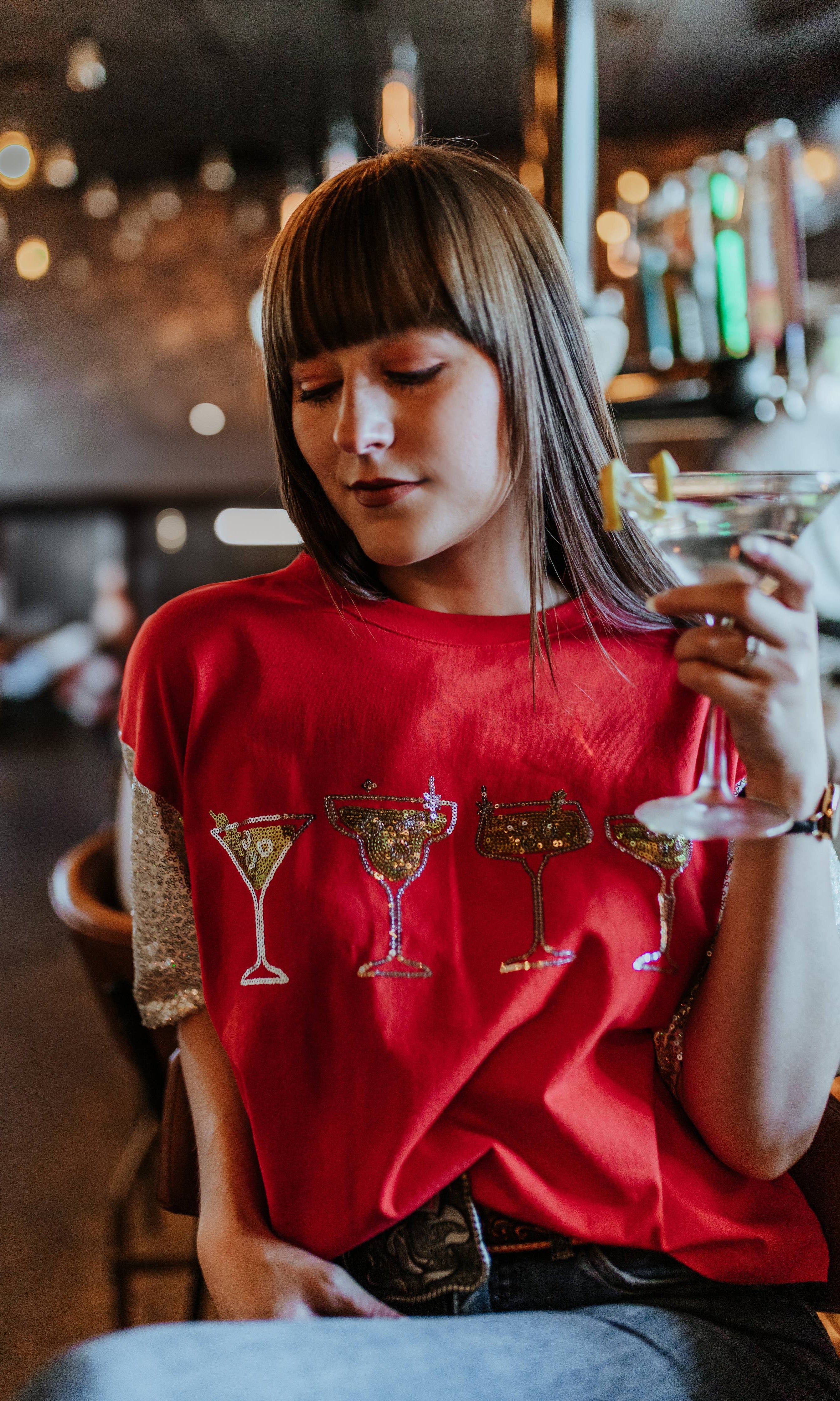 Woman in a red t-shirt with cocktail graphics holding a martini glass in a bar setting.
