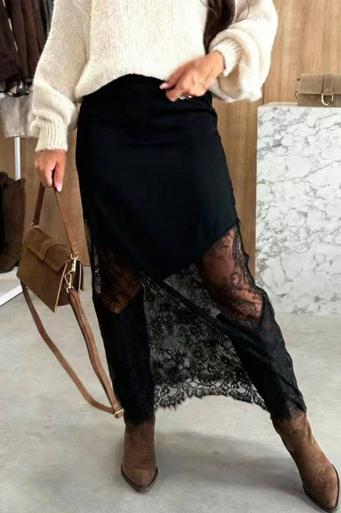 Person wearing a black lace skirt with a white sweater and brown boots, holding a brown handbag.