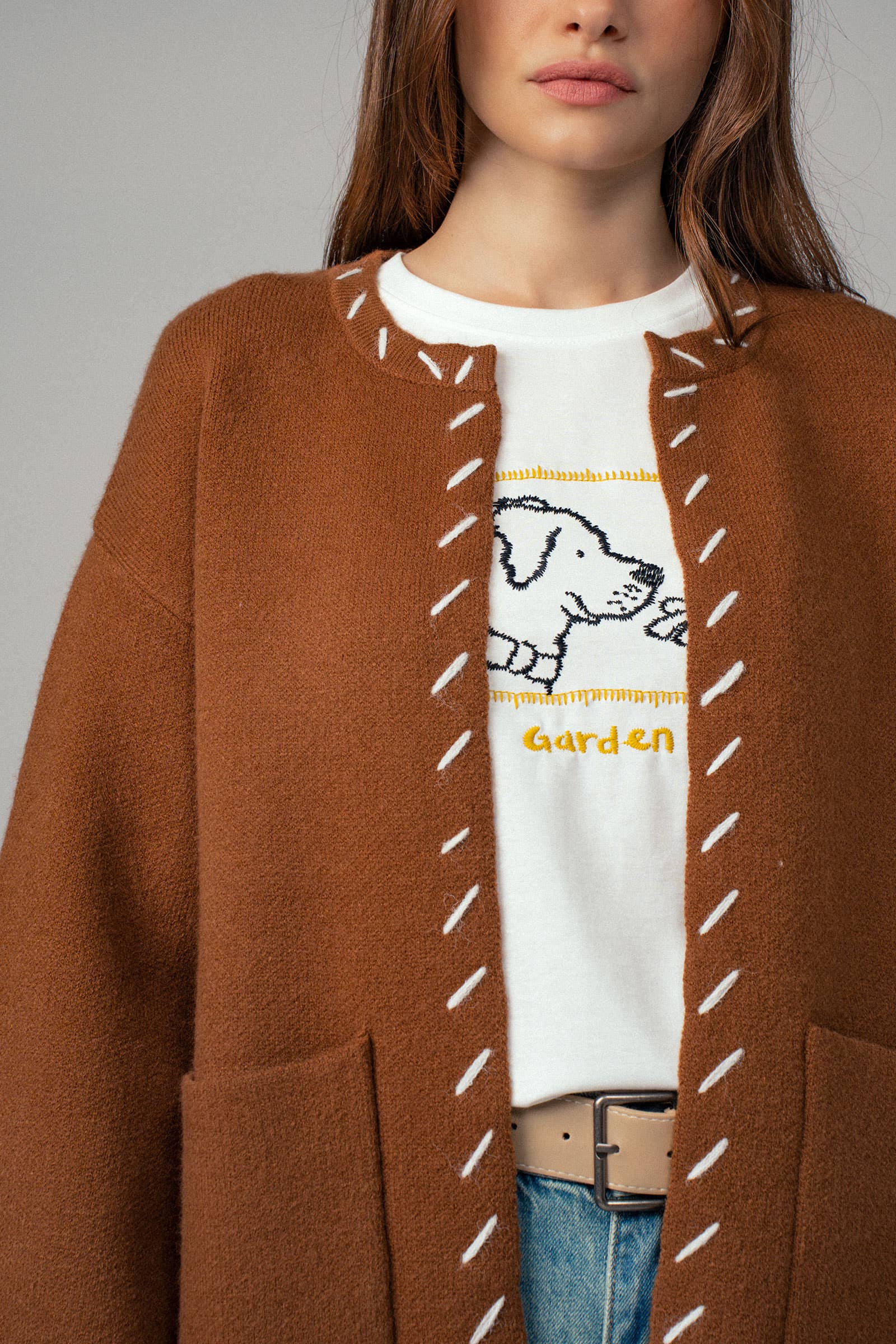 Close up of woman wearing a brown open coat with white bold stitching trim on a gray