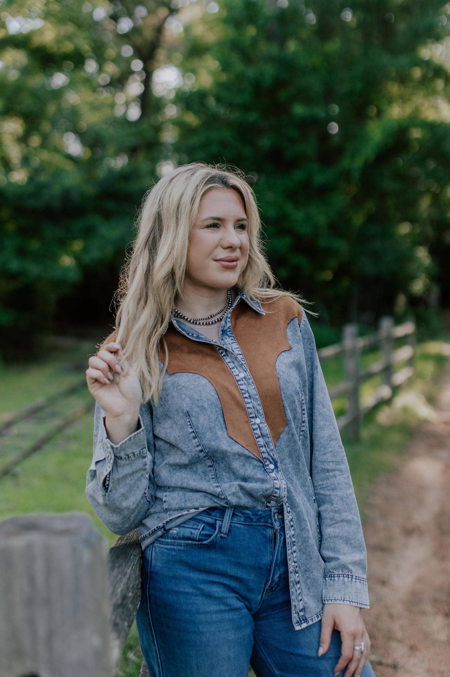 Woman wearing a denim and brown shirt and blue jeans outdoors with greenery in the background