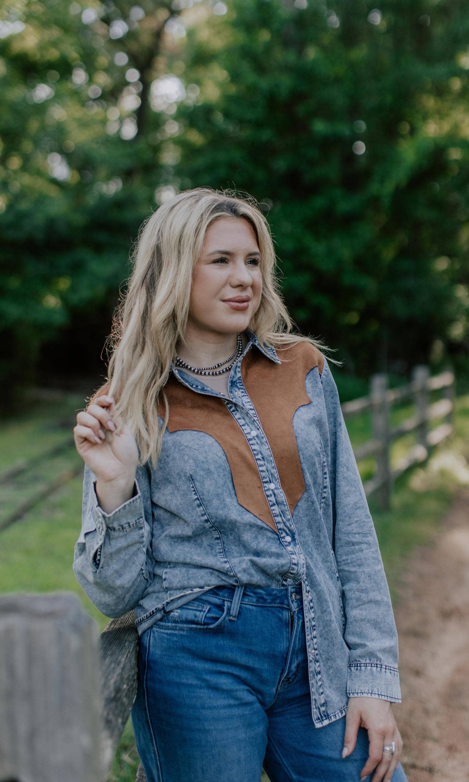 Woman wearing a denim and brown shirt and blue jeans outdoors with greenery in the background