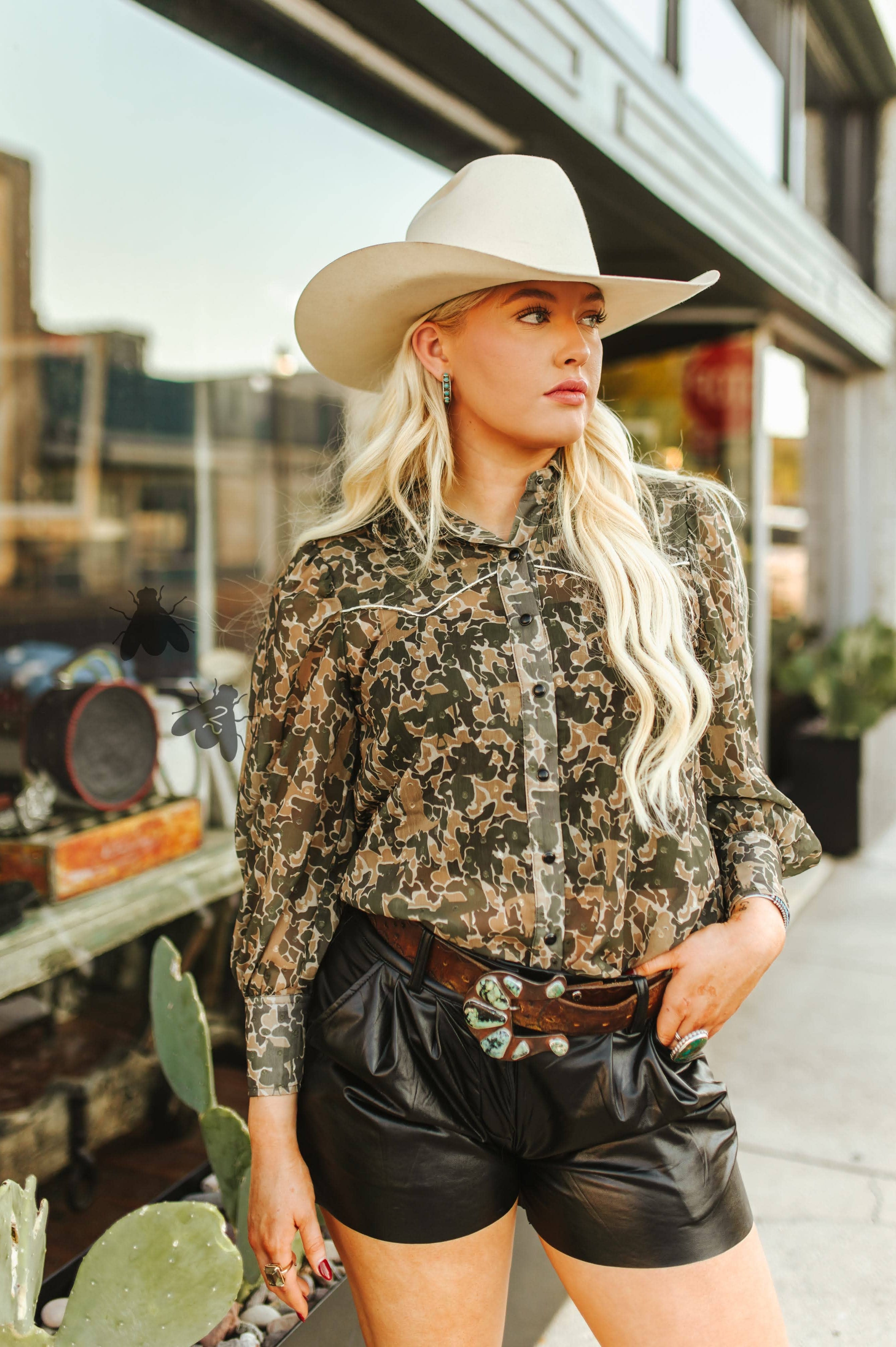 Woman wearing a camouflage shirt, leather shorts, and a cowboy hat in an urban setting.