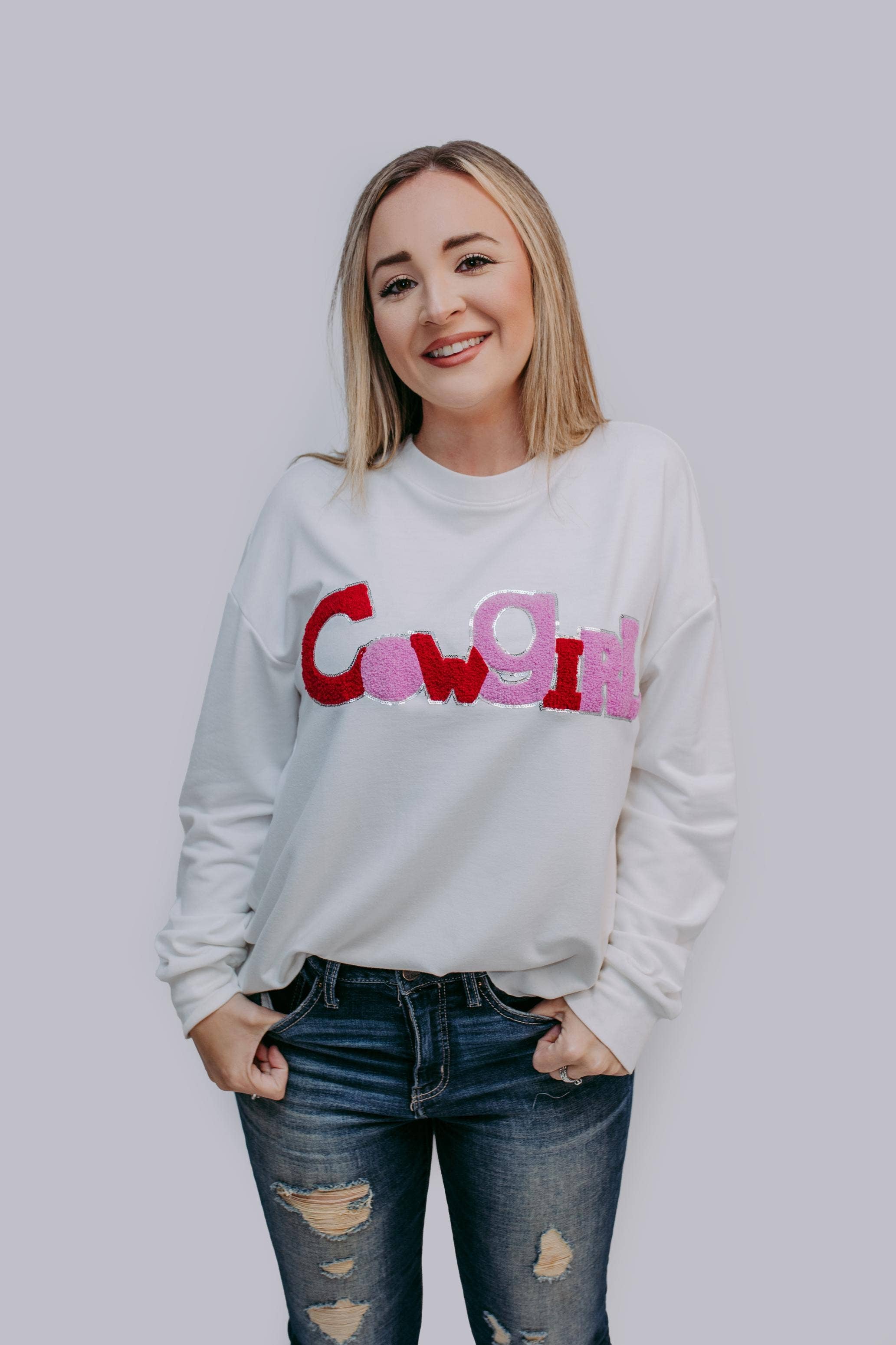 Woman wearing a white sweatshirt with colorful text"COWGIRL" on a plain background