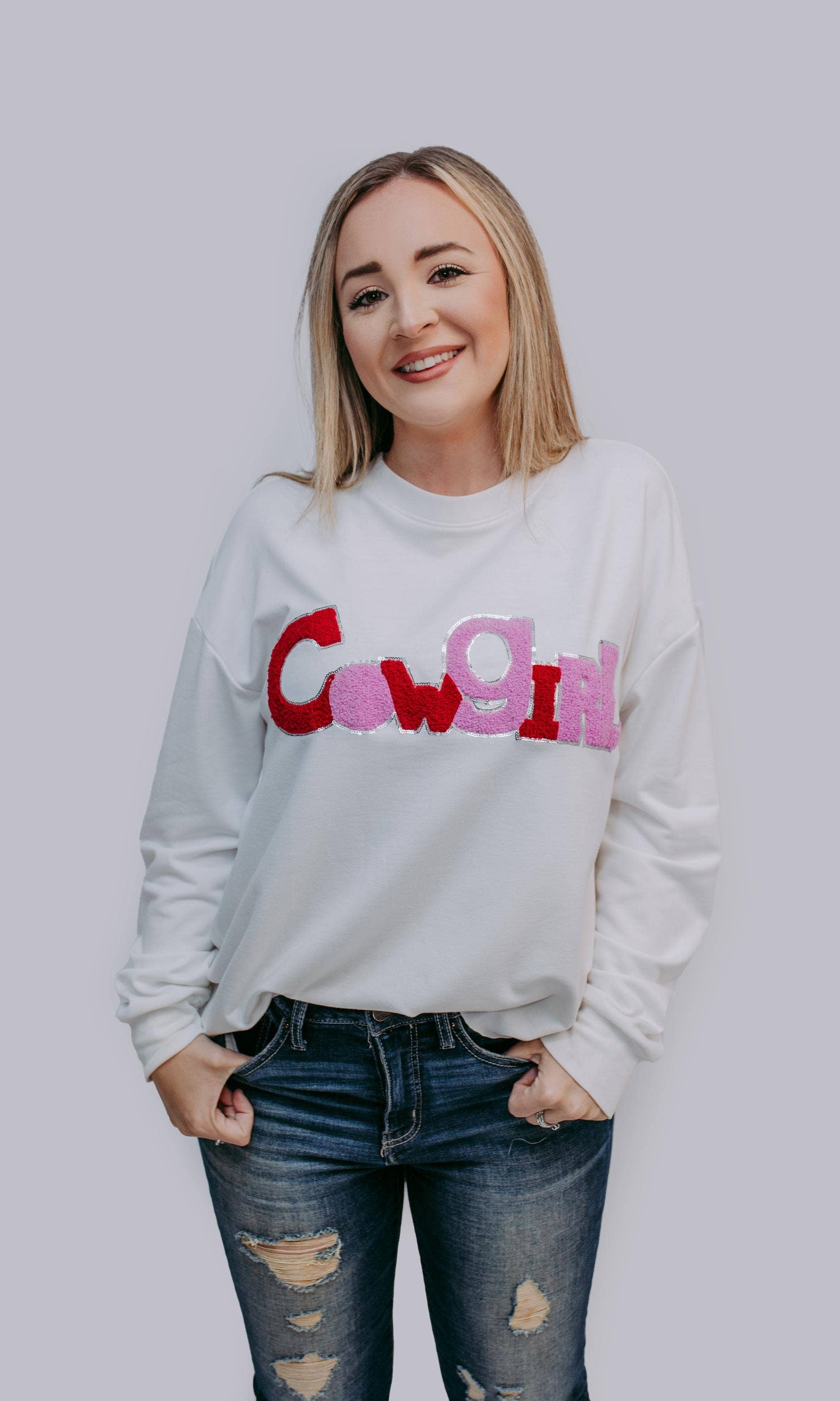 Woman wearing a white sweatshirt with colorful text"COWGIRL" on a plain background