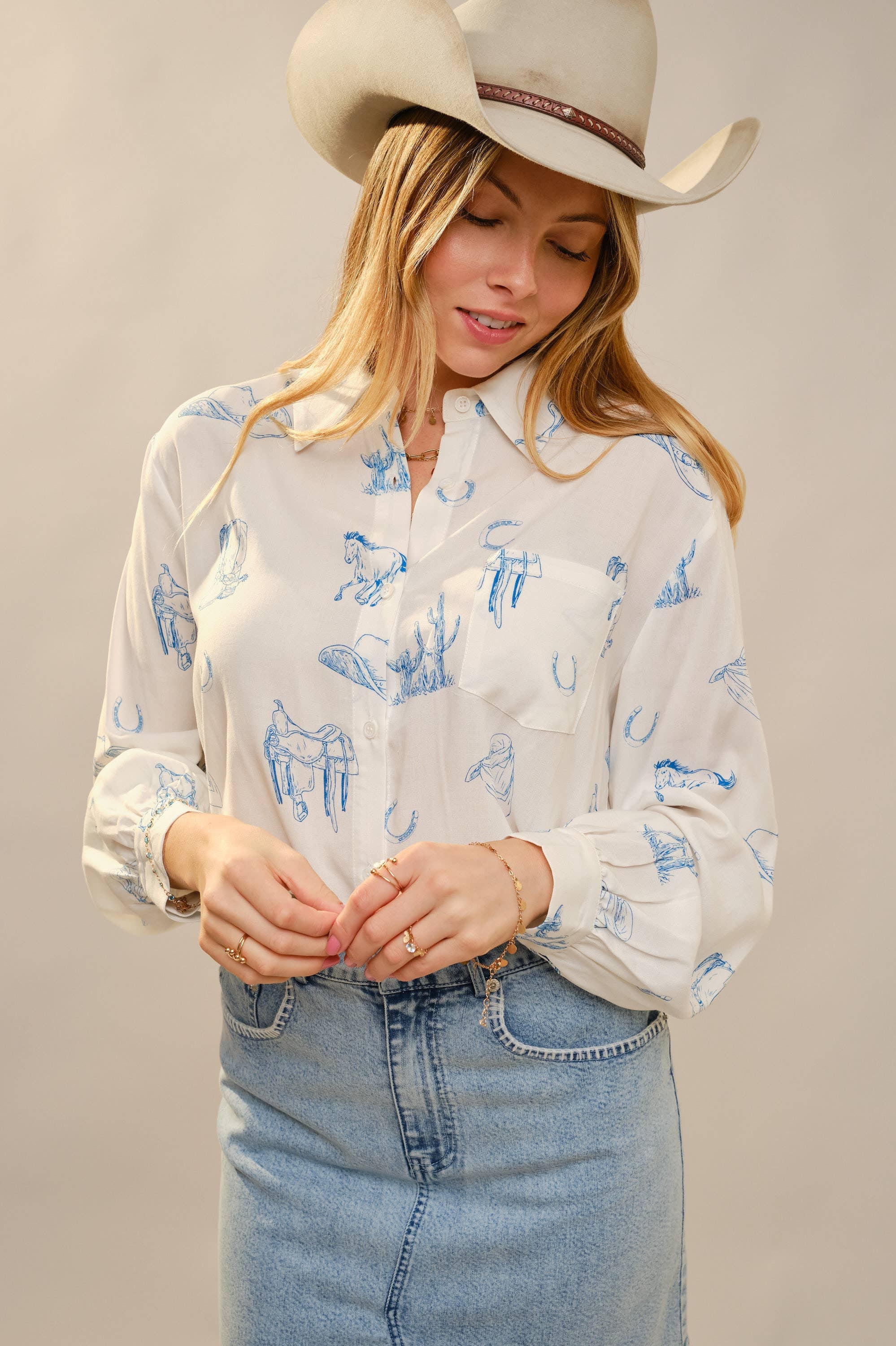 Woman wearing a white blouse with blue patterns, denim skirt, and cowboy hat against a plain background