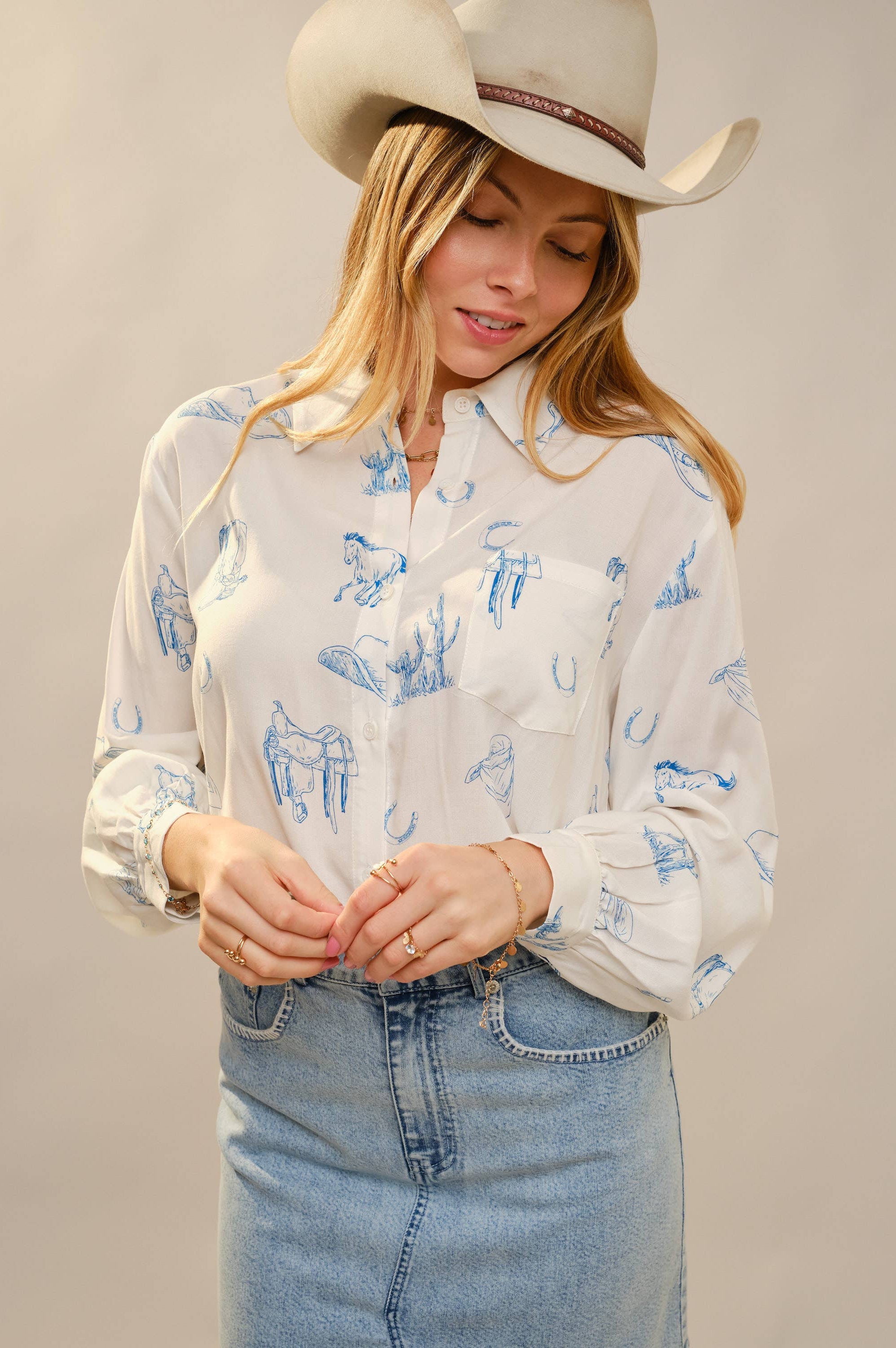 Woman wearing a white blouse with blue patterns, denim skirt, and cowboy hat against a plain background