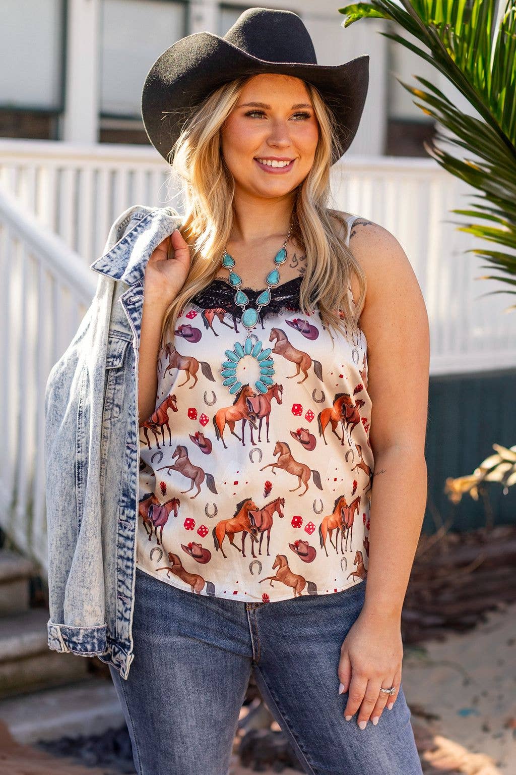 Woman wearing a sleeveless top with horse print, jeans, and a cowboy hat outdoors.