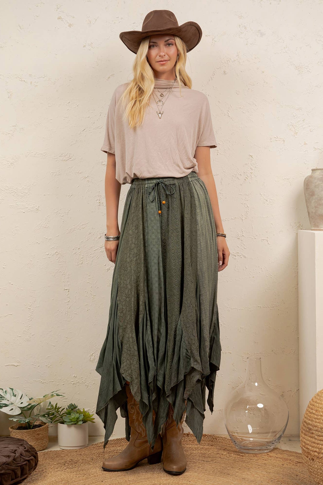 A woman stands indoors wearing a brown cowboy hat, a light beige short-sleeve top, a dark olive green high-low skirt with button details, and brown leather boots. The setting includes a textured cream wall, a white pedestal with dried grass in a vase, and woven poufs.
