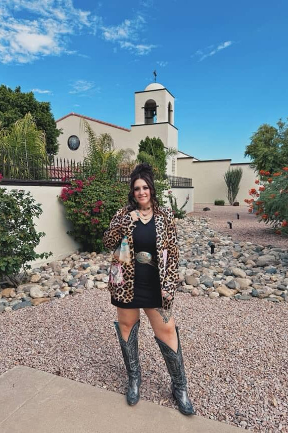Woman in leopard print jacket and black dress standing in front of a church with a bell tower.