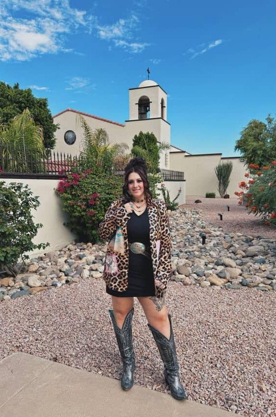 Woman in leopard print jacket and black dress standing in front of a church with a bell tower.