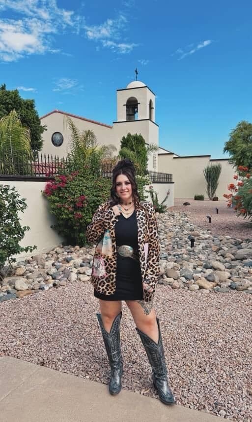 Woman in leopard print jacket and black dress standing in front of a church with a bell tower.