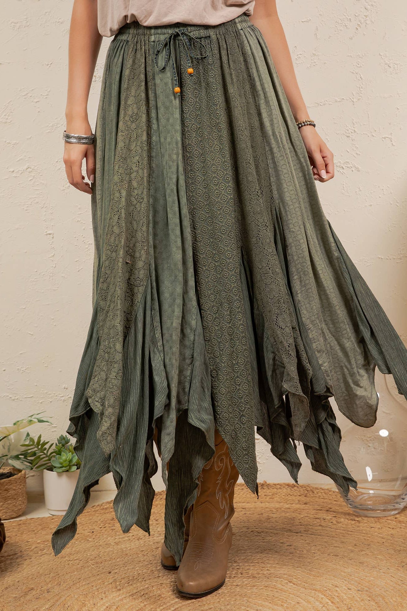 A woman wearing a long, flowing sage green skirt with a tie waist and brown ankle boots, standing in a styled room with plants and woven decor.