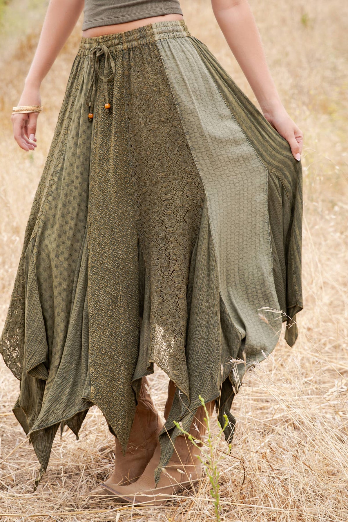 An olive green bohemian maxi skirt with mixed lace and textured fabric panels, worn by a model standing in a dry grass field.
