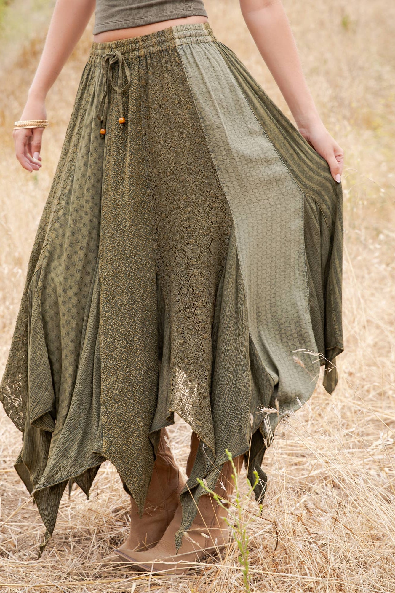 An olive green bohemian maxi skirt with mixed lace and textured fabric panels, worn by a model standing in a dry grass field.