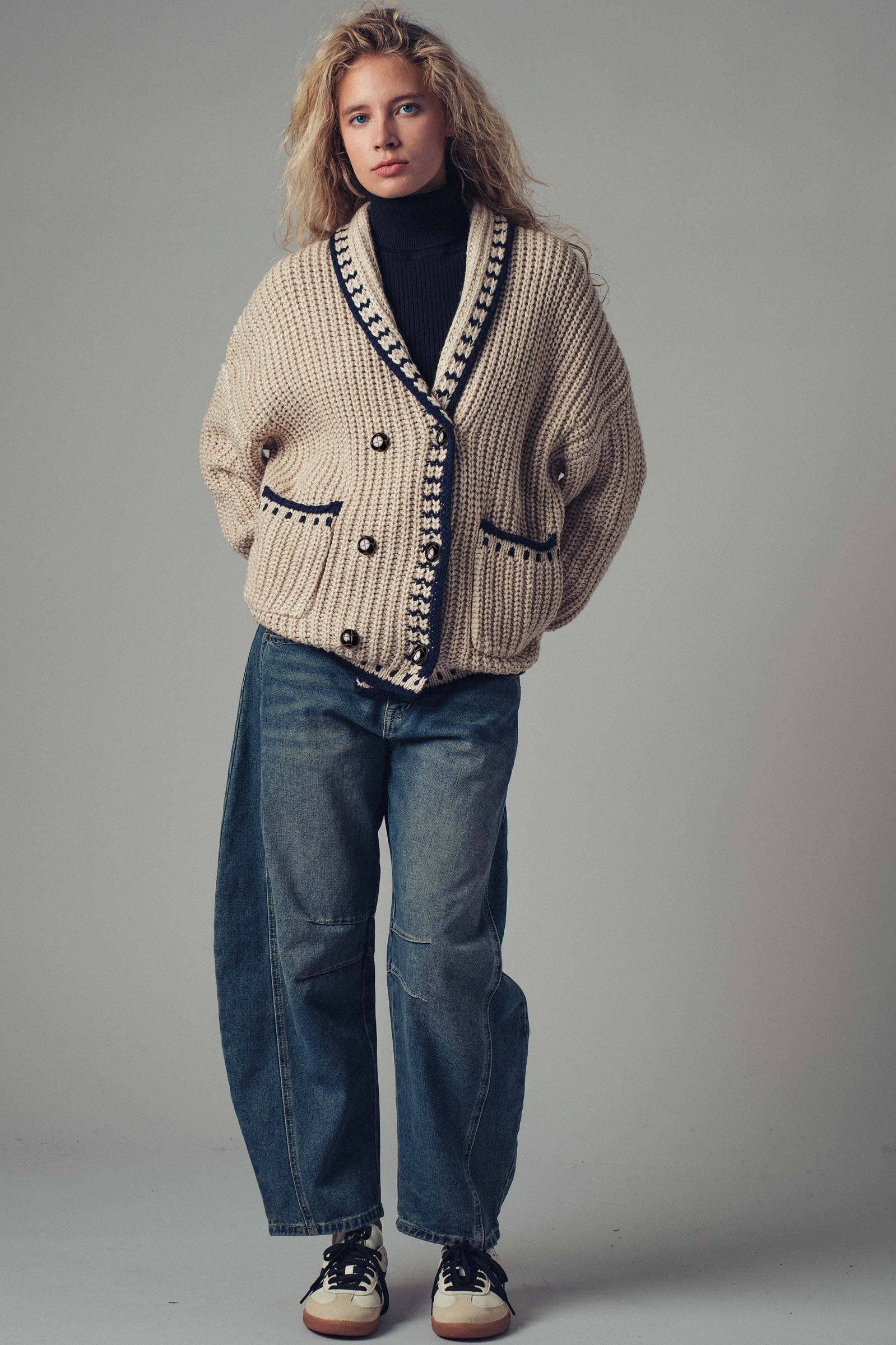 Woman wearing a beige cardigan with black trim and blue jeans on a gray background