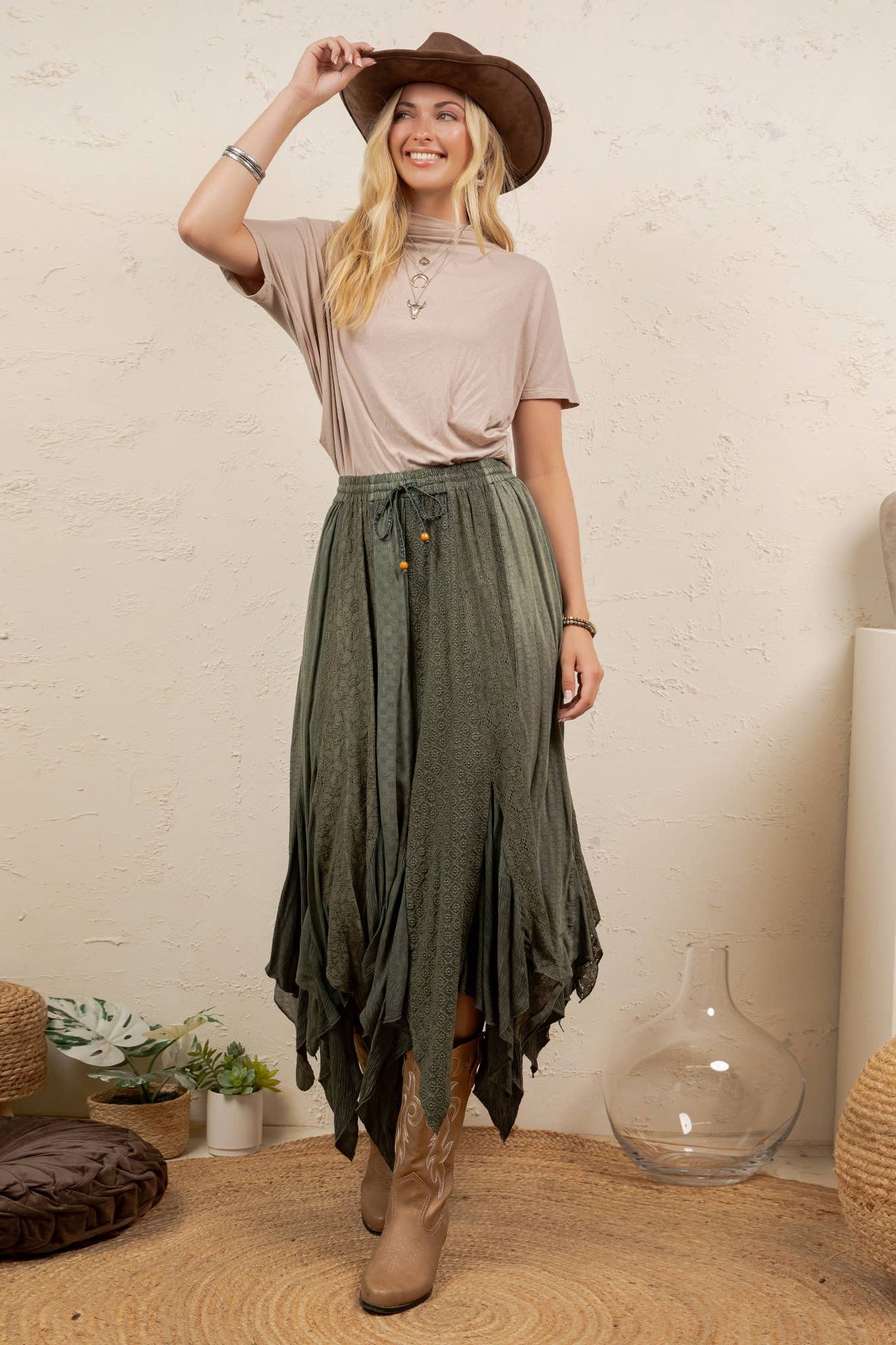 A woman stands in a styled indoor setting wearing a brown wide-brim hat, a light beige short-sleeve top, and a dark olive green high-waisted, tiered maxi skirt with brown leather boots.