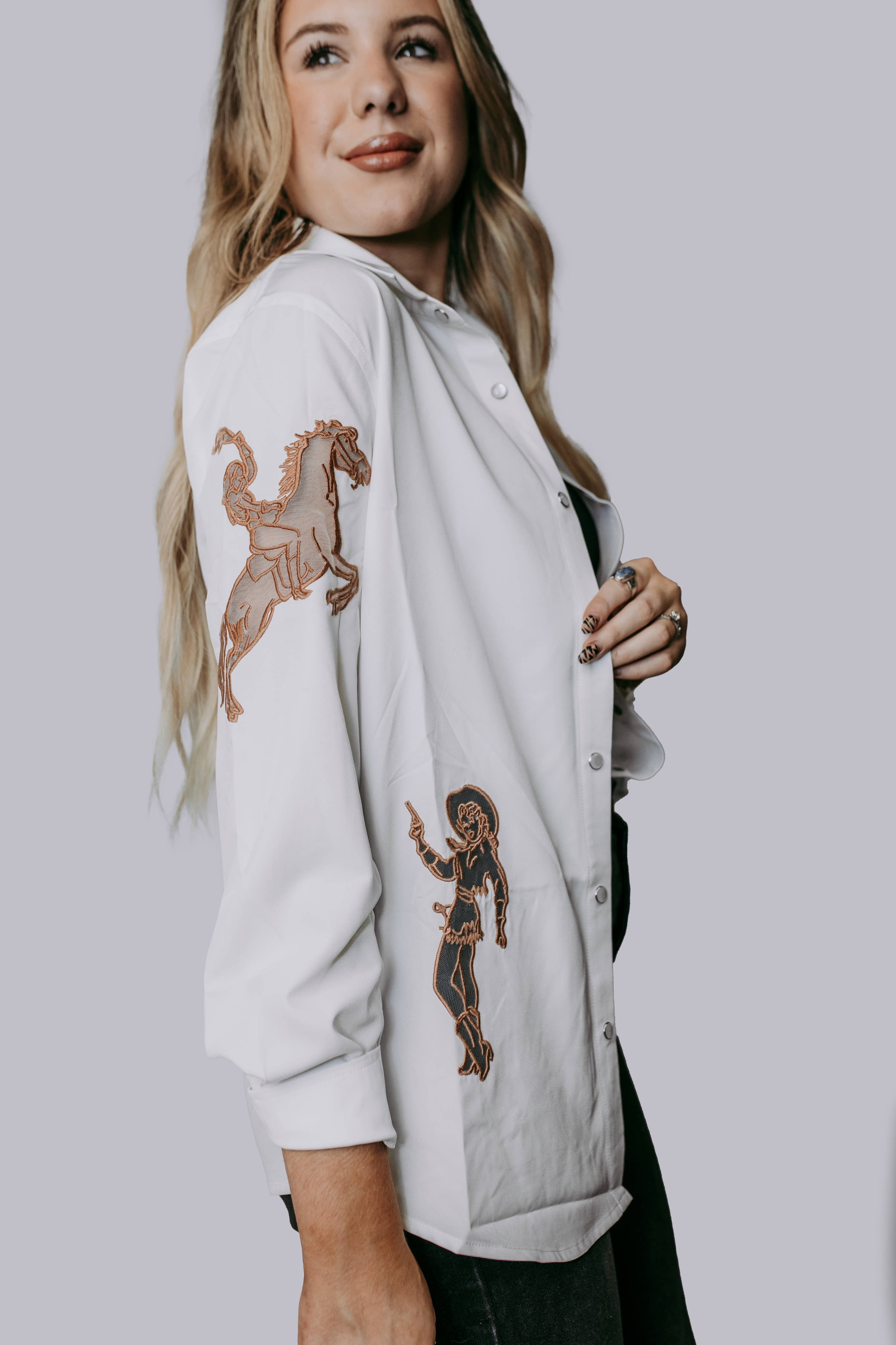 A woman smiling and posing in a white button-down blouse with cut out embroidery western horseshoes and cowboys with pearl snap buttons, paired with dark pants.