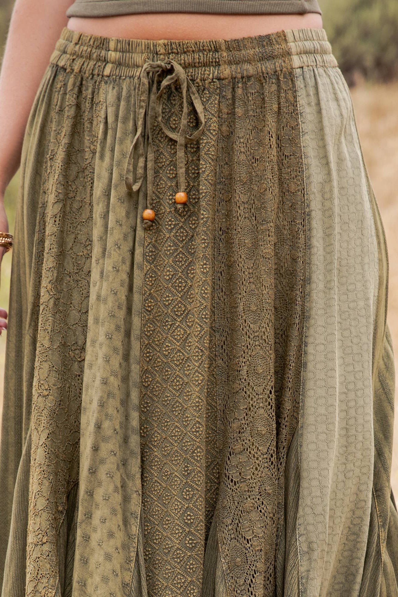 A close-up of a person wearing a loose, olive green patterned skirt with an elastic drawstring waistband, standing outdoors against a blurred natural background.