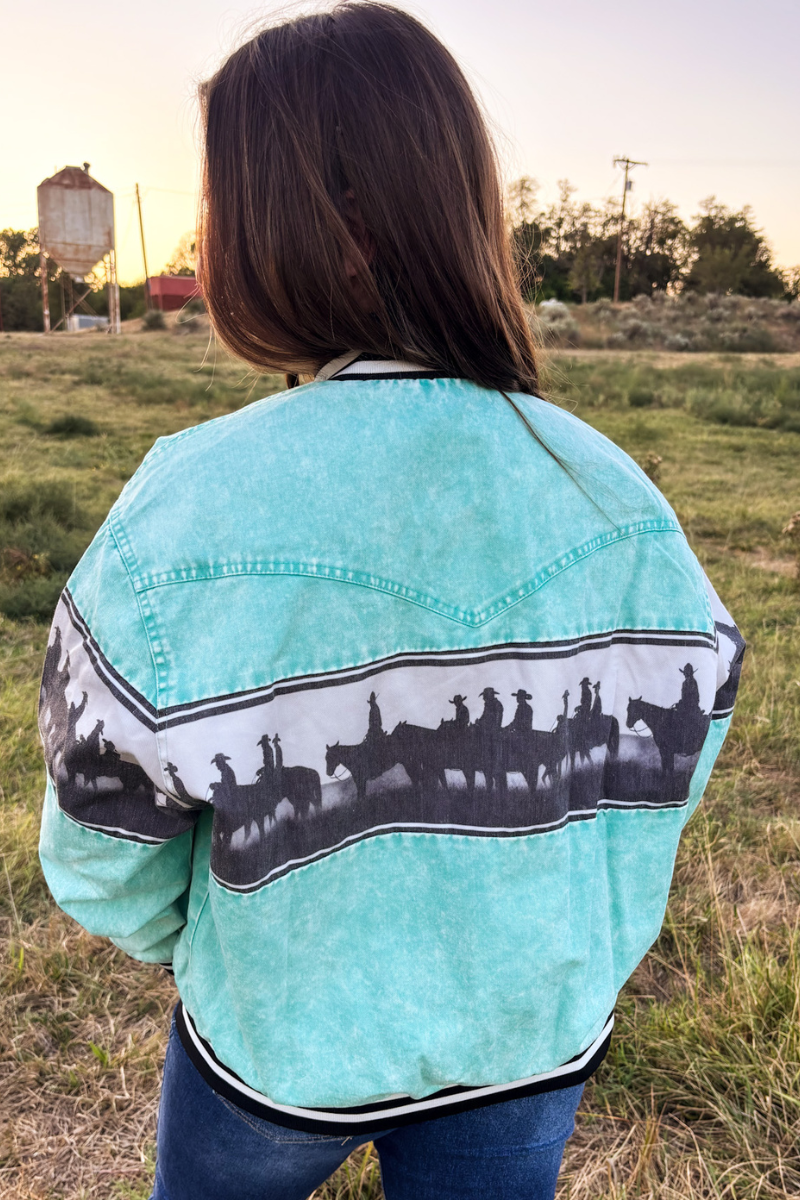 A person viewed from behind wearing a turquoise cropped jacket with a black and white western rider graphic across the back, standing in an outdoor grassy field at sunset.