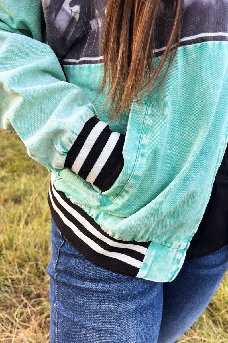 A close-up of a person wearing a mint green and black color-blocked jacket with black and white striped ribbed cuffs and hem, paired with blue jeans, standing outdoors on grass.