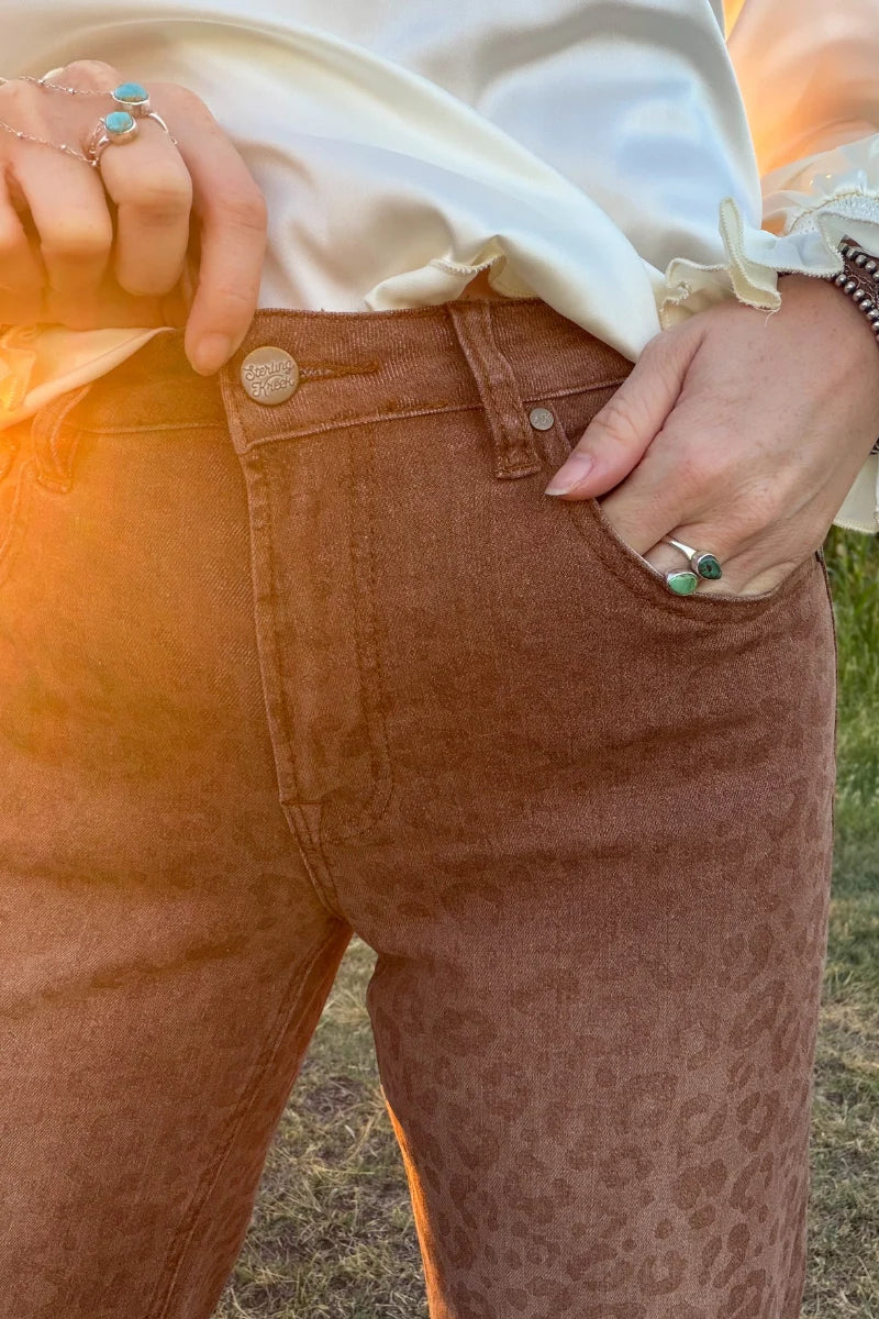 Close-up of Sand Cat Cuffed Jeans showing the tonal leopard print, high-rise waistband, front pocket, button closure, and warm camel-brown wash.