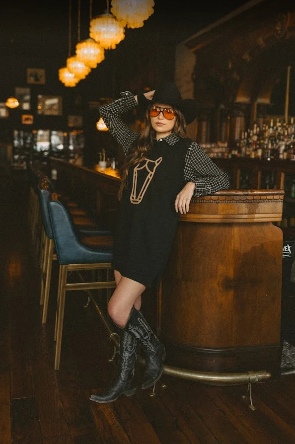 A woman posing in a rustic bar setting wearing a black cowgirl-style dress with a horse graphic, a black-and-white checkered long-sleeve shirt, a black cowboy hat, and black cowboy boots.