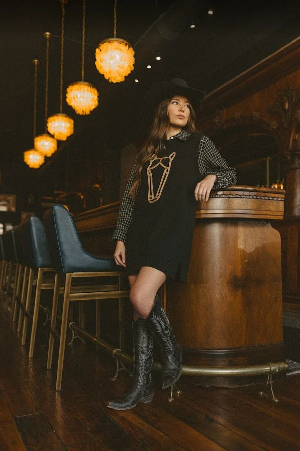 A woman wearing a black cowboy-style mini dress with a gold horse outline, a black-and-white gingham shirt, a black cowboy hat, and black leather boots, leaning against a wooden bar in a dimly lit bar setting.
