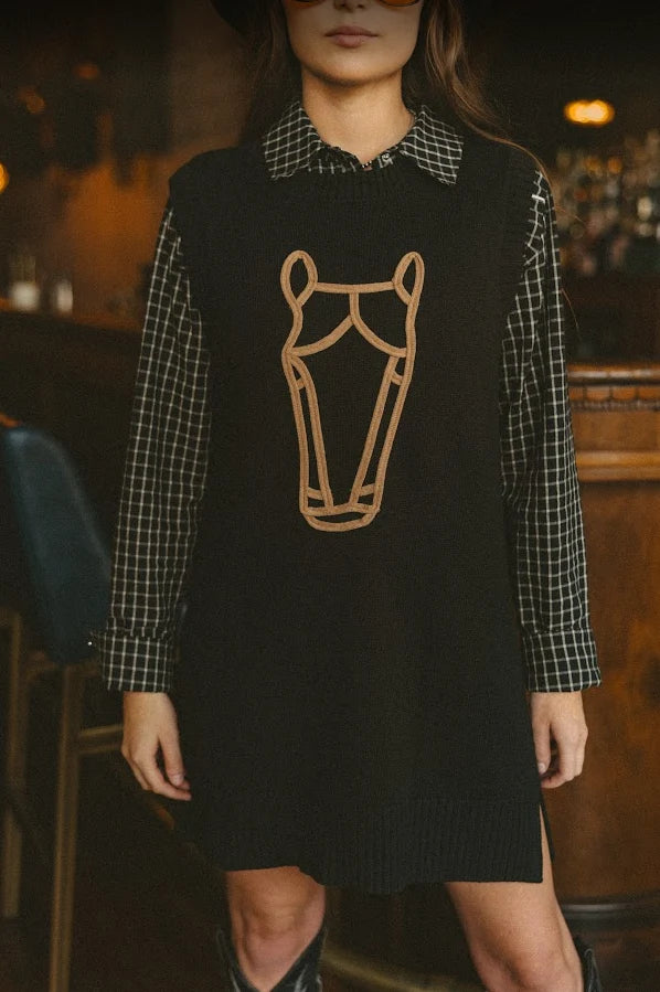 A woman wearing a black knit sweater vest over a black-and-white checkered long-sleeve shirt, with a tan embroidered horse head outline on the front, photographed in a dimly lit indoor setting.