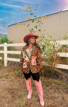 Woman in a leopard print jacket and pink cowboy boots standing in a field with sunflowers and a white fence in the background.