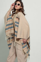 Woman wearing a beige poncho with a  Tan background and light blue, brown, light rust, and grey aztec pattern design  in stripes with tassel trim along bottom hem. Light weight fabric 