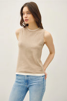 Woman wearing a tan sleeveless top with lighter contrasting band at the bottom of the tops and blue jeans on a plain background