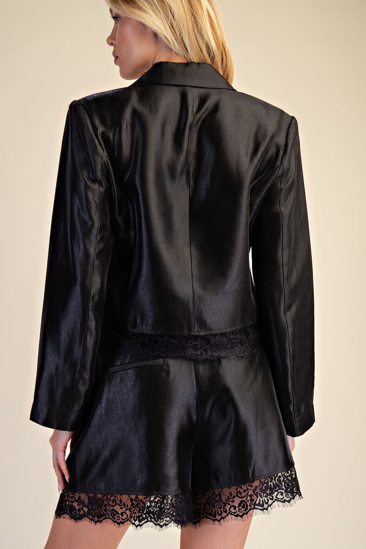 A model shown from the back wearing a black satin blazer and matching shorts with black lace trim, set against a solid beige background.