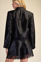 A model shown from the back wearing a black satin blazer and matching shorts with black lace trim, set against a solid beige background.