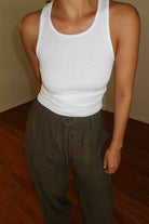 Person wearing a white tank top and brown elastic waist pants on a wooden floor.