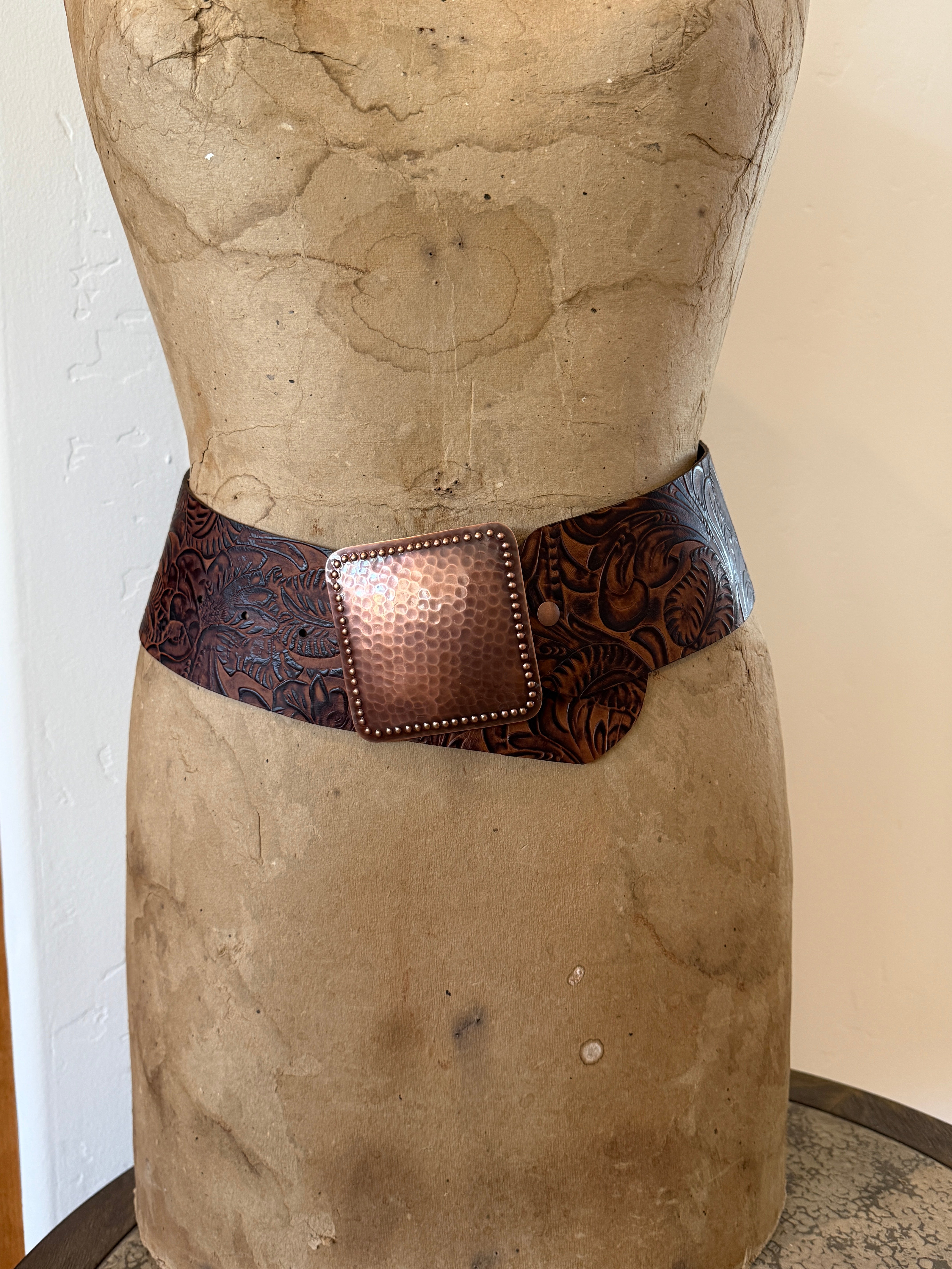A wide brown tooled leather belt with a hammered copper-toned rectangular buckle, displayed on a vintage tan dress form mannequin.