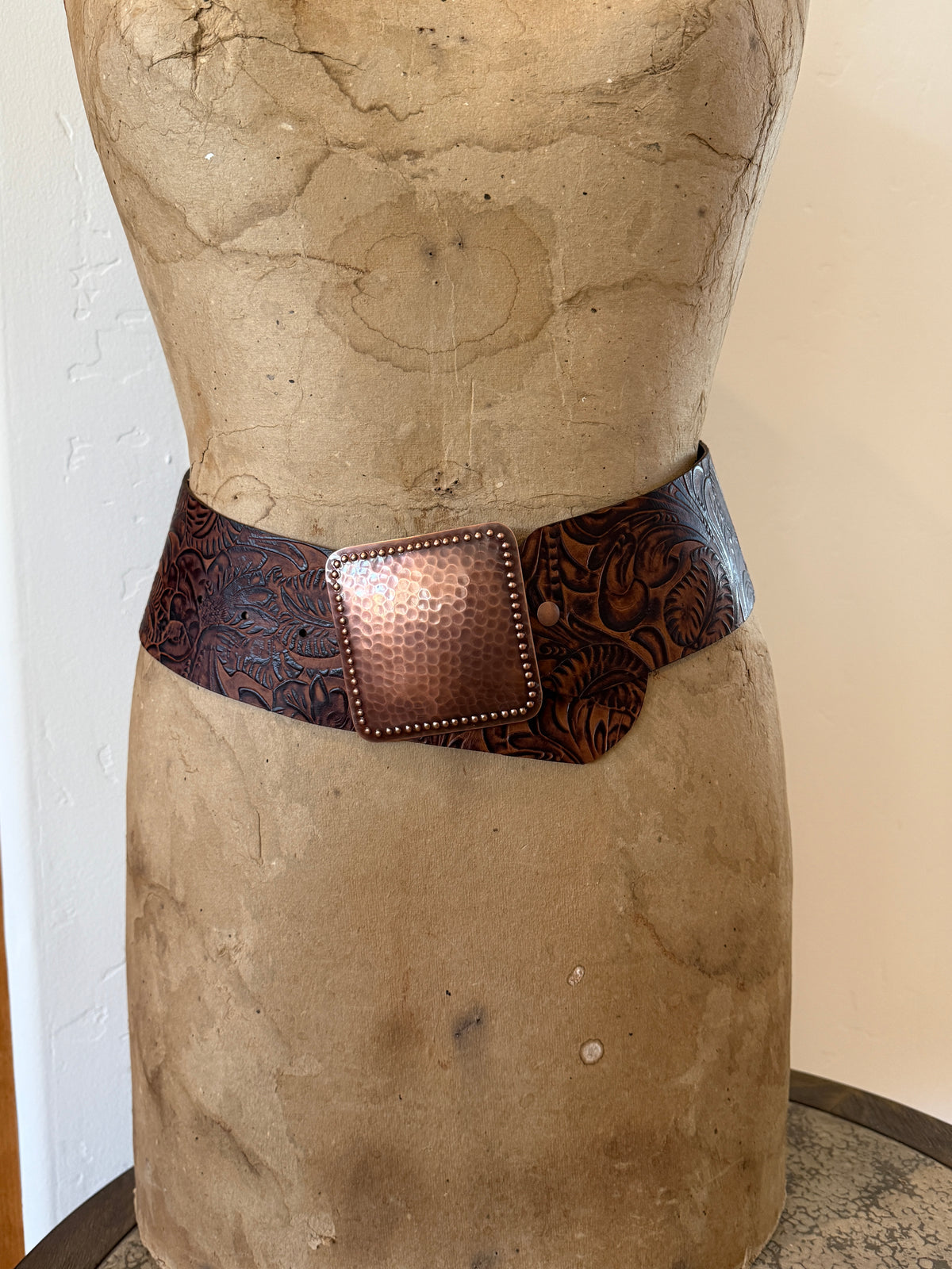 A wide brown tooled leather belt with a hammered copper-toned rectangular buckle, displayed on a vintage tan dress form mannequin.