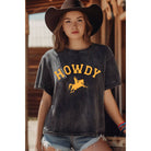 A young woman wearing a dark gray acid-wash t-shirt with a yellow 'HOWDY' cowboy graphic, paired with a black cowboy hat and denim shorts, posed in front of a rustic wooden barn background.